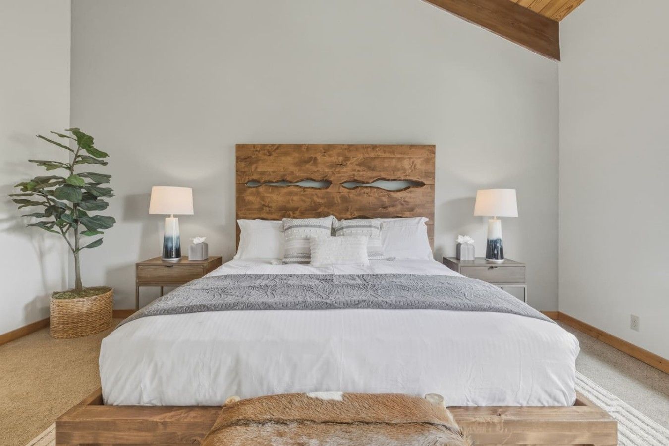 A bedroom with a wooden headboard, a large bed with white bedding, two bedside tables with lamps, and a potted plant.