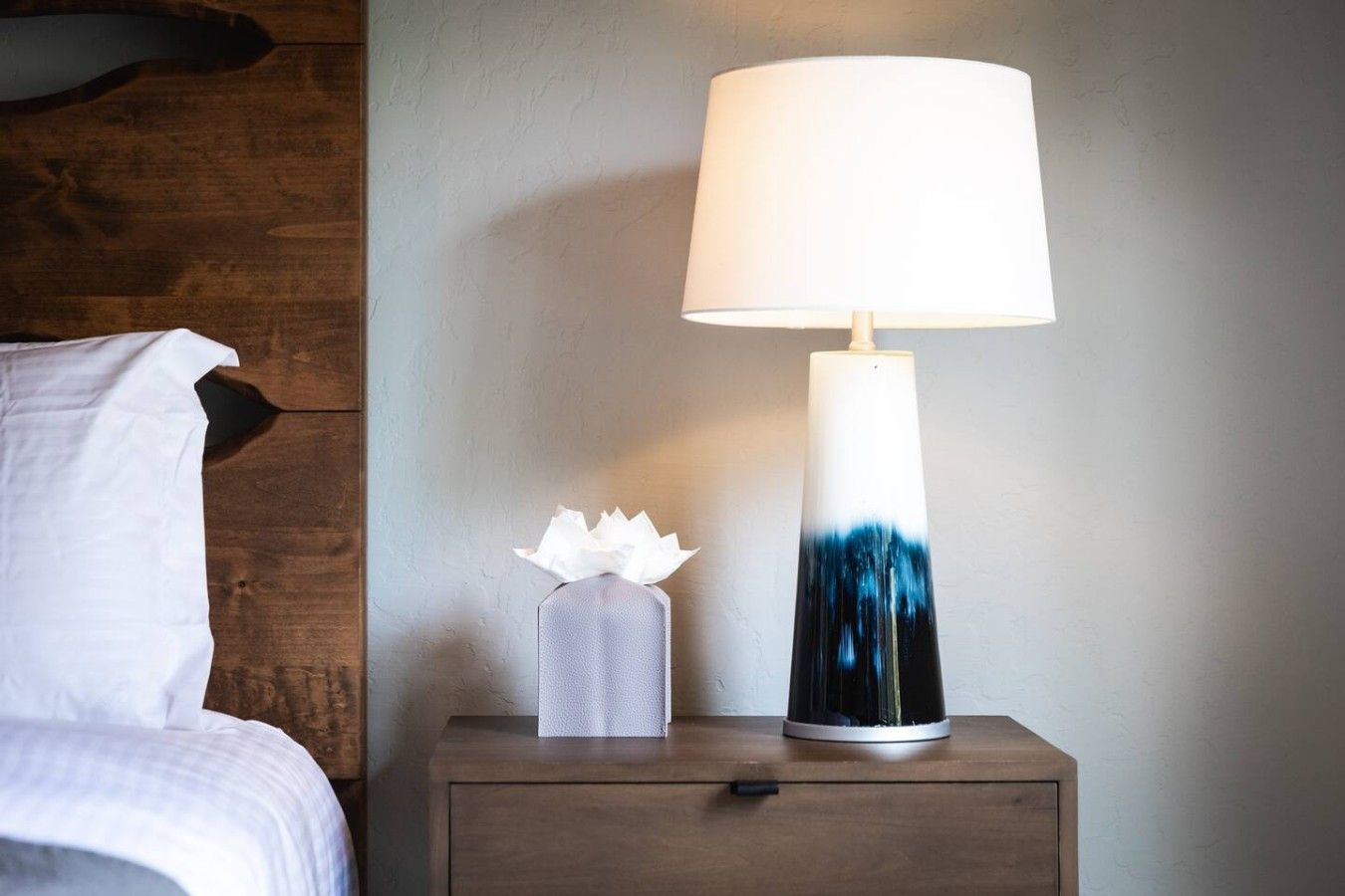 A bedside table with a white and blue gradient lamp, a tissue box, and a white pillow on a wooden nightstand.