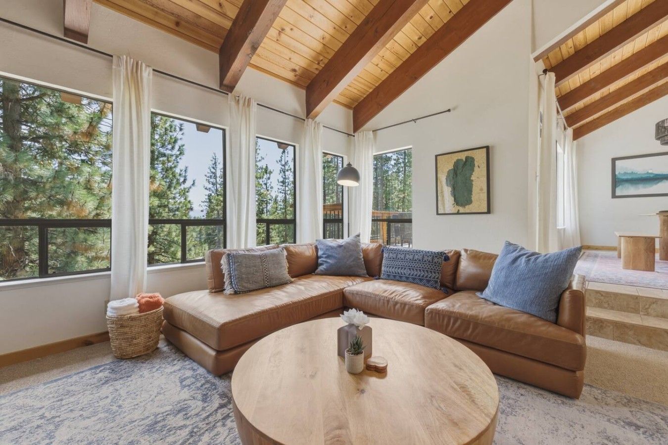 A spacious living room with a large L-shaped sectional sofa, a round wooden coffee table, and large windows offering a view of trees.