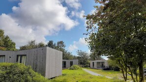 A modern building with a flat roof and large windows, surrounded by greenery and trees. - Large modern holiday home with pool close to the beach (Groede)