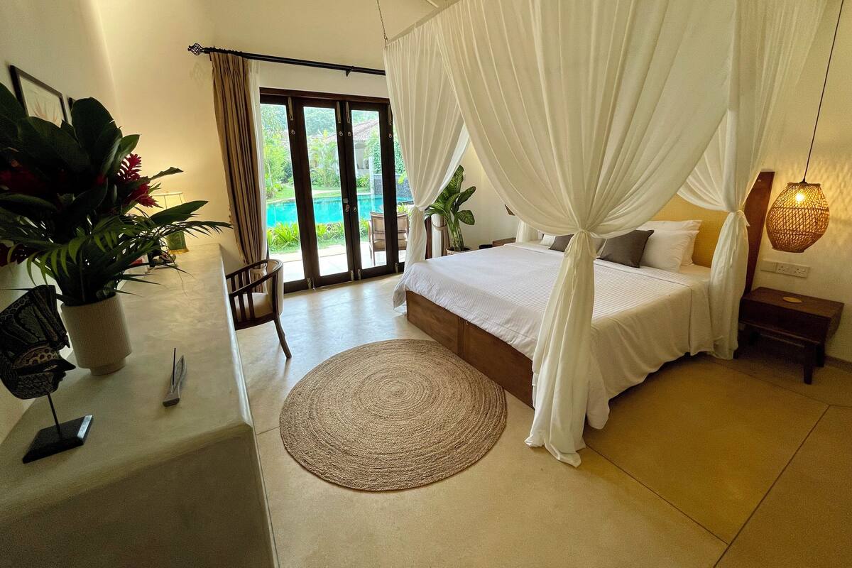 A bedroom with a bed, a bedside table, a nightstand, a potted plant, a chair, and a view of a pool through a glass door.