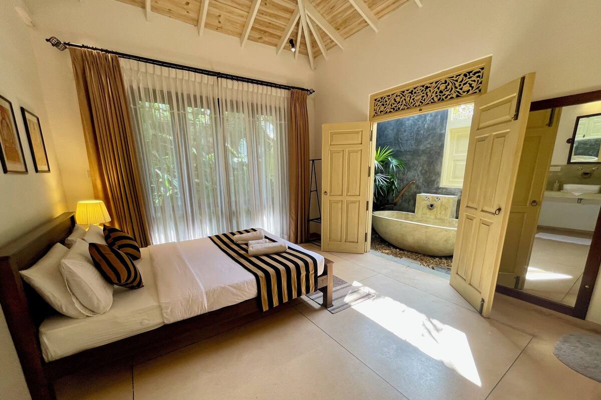 A bedroom with a bed, curtains, a bedside lamp, and a view of a bathroom with a bathtub.