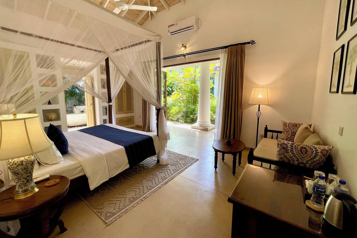 A bedroom with a canopy bed, a bedside table with a lamp, a small table with a lamp, and a view of greenery outside.
