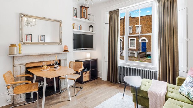 A living room with a fireplace, a dining table with chairs, a green sofa, a TV, and a view of a brick building through a window. - The Barnsbury, London Apartment w/WIFI (London)