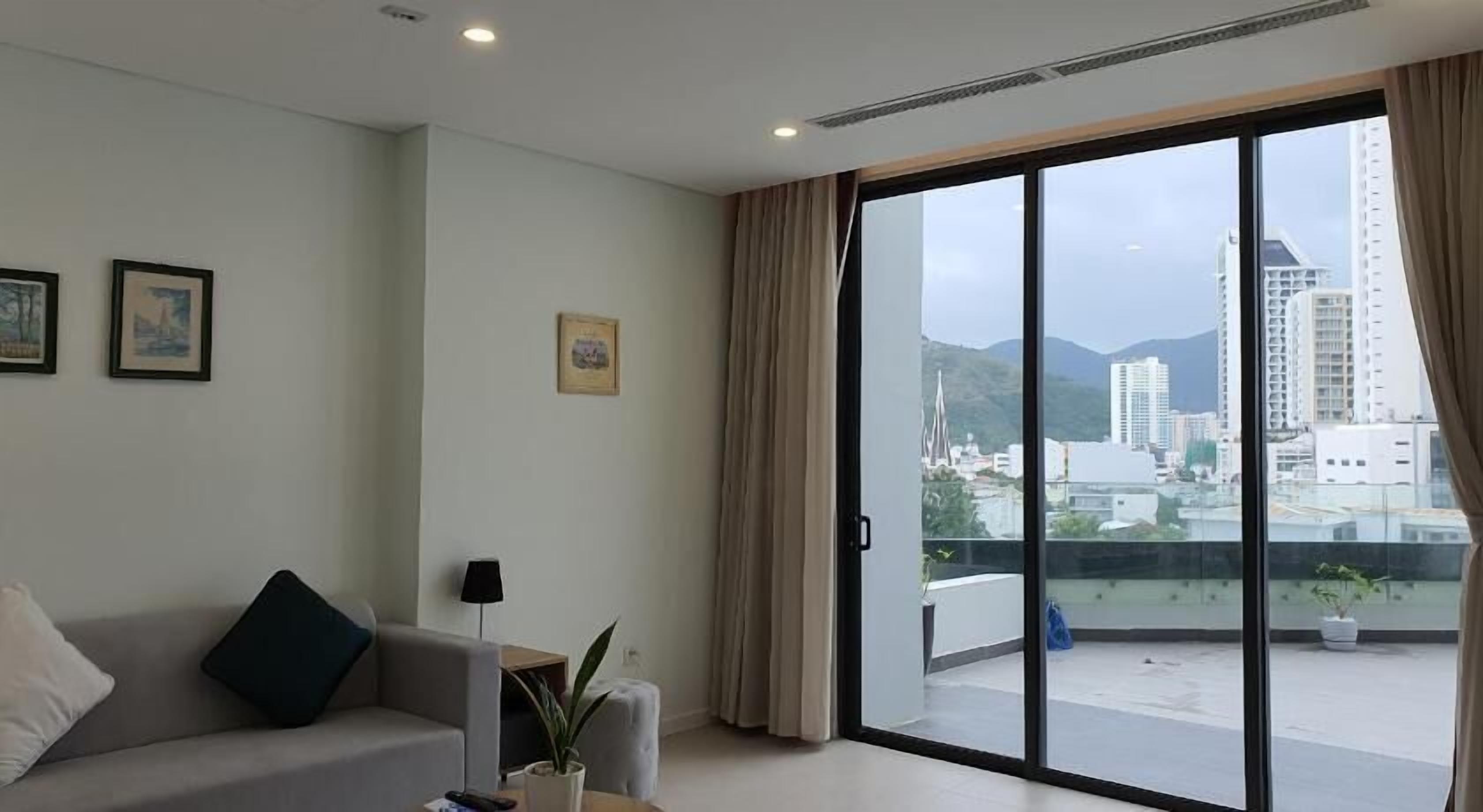 Ein modernes Wohnzimmer mit einer Couch, einem kleinen Tisch und einem Blumentopf. Eine Glastür öffnet sich zu einem Balkon mit Blick auf die Stadt.