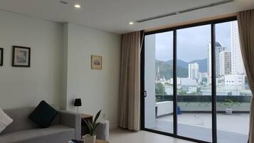 Ein modernes Wohnzimmer mit einer Couch, einem kleinen Tisch und einem Blumentopf. Eine Glastür öffnet sich zu einem Balkon mit Blick auf die Stadt.