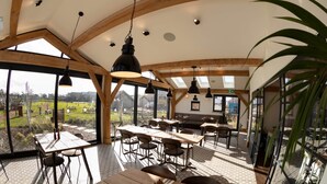 Un comedor amplio con techos altos y ventanas grandes que ofrecen vistas al exterior. Mesas y sillas están ordenadas, y lámparas colgantes adornan el techo.