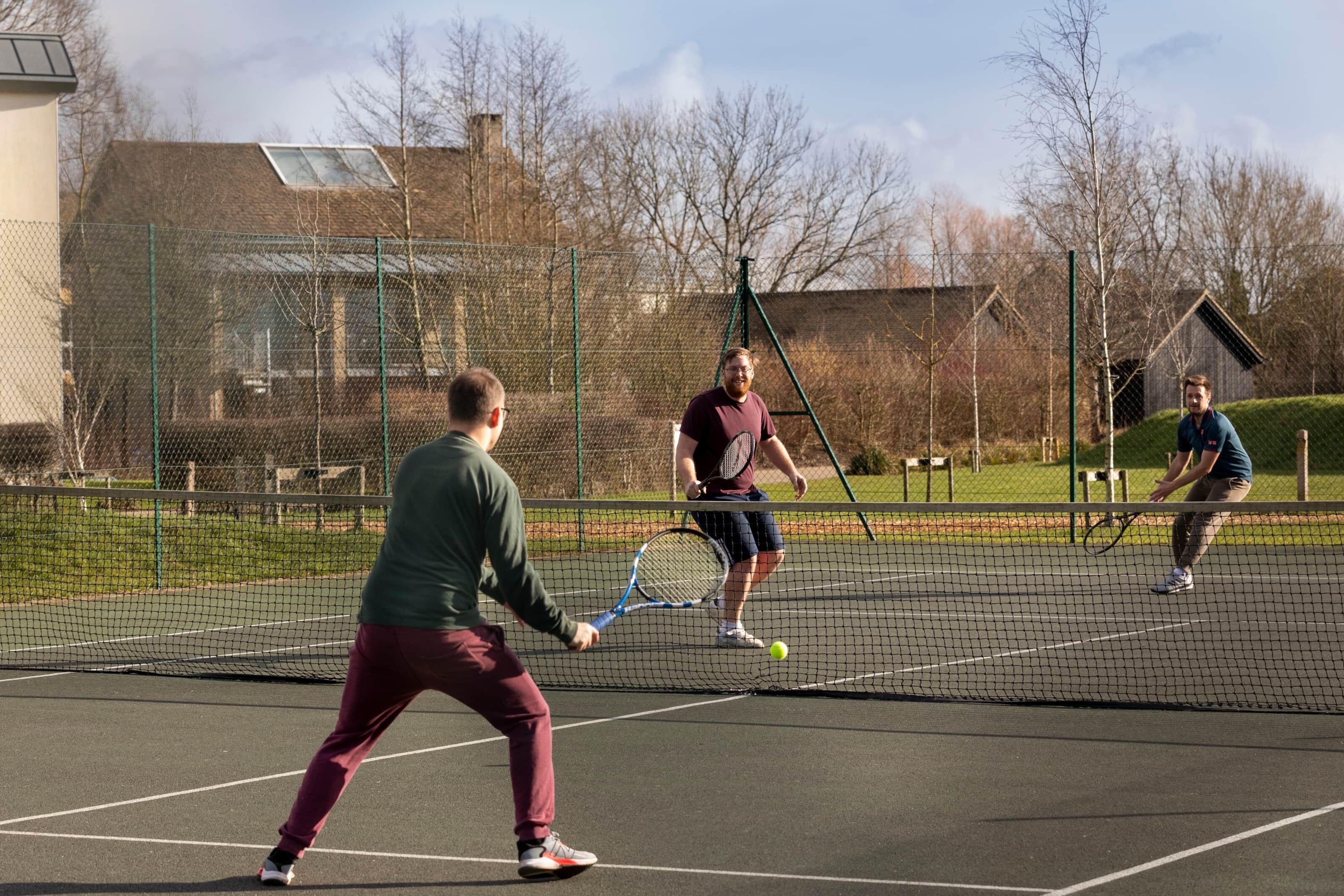 A tennis court with a net, surrounded by a fence, and buildings in the background.