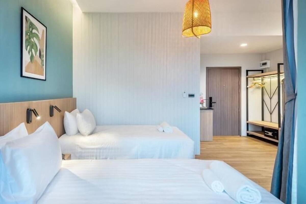 A hotel room with two beds, a wooden headboard, a wall-mounted picture, a hanging light fixture, and a visible bathroom area with a mirror and towel rack.
