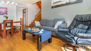 Un salon avec un canapé gris, un fauteuil en cuir noir, une table à manger en bois avec des chaises et une nappe bleue. On aperçoit un escalier au fond et un tableau accroché au mur.