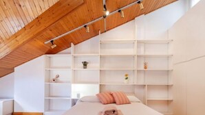 Une chambre avec un plafond en pente, des poutres en bois, un lit avec un oreiller rose et une serviette pliée, des étagères blanches et une petite plante en pot.