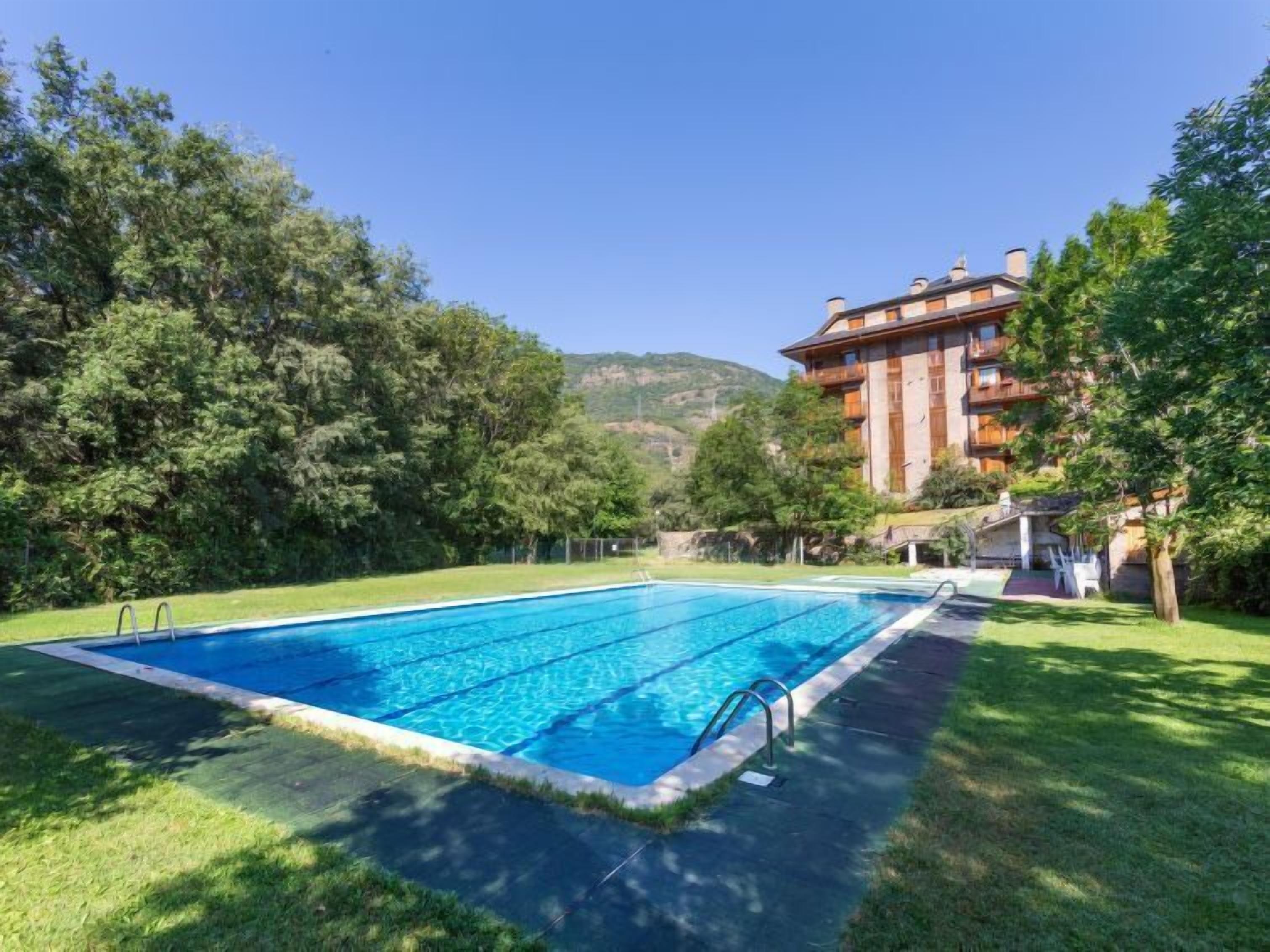 Uma piscina retangular com água azul cristalina, rodeada por relva bem cuidada e árvores.