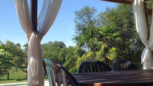 An outdoor pool area with a wooden deck, chairs, and a pergola with white curtains.