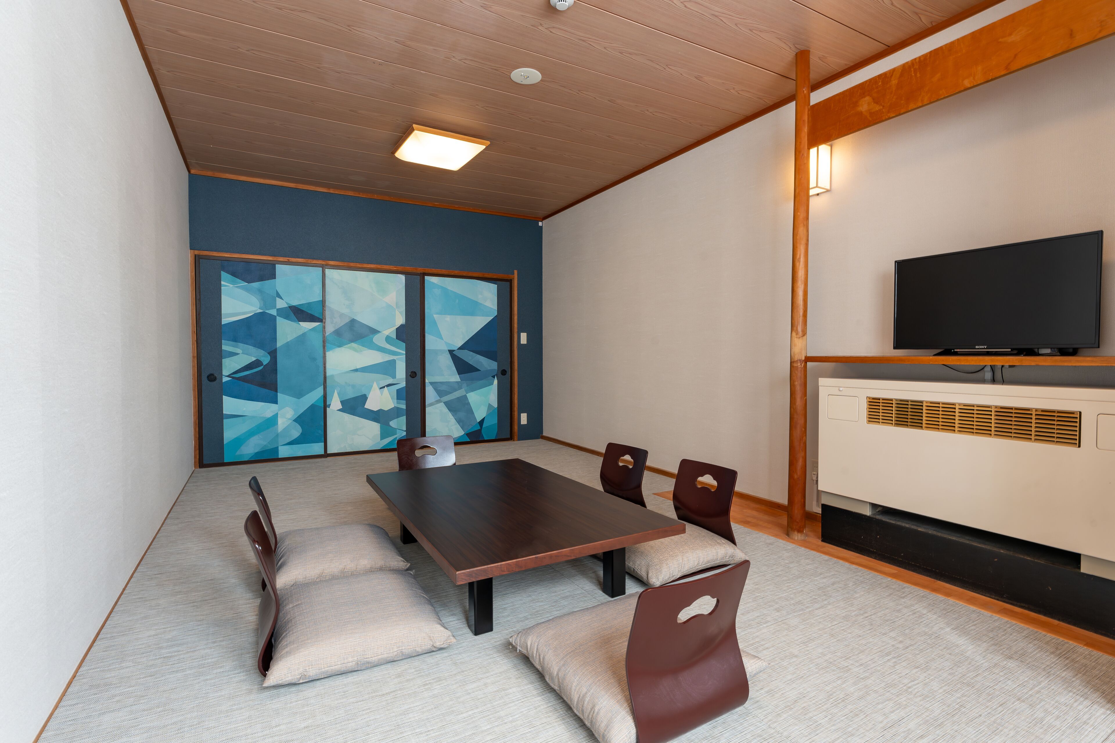 Een moderne kamer met een houten plafond, een grote tafel met stoelen, een flatscreen televisie en een schuifdeur met abstracte kunst.