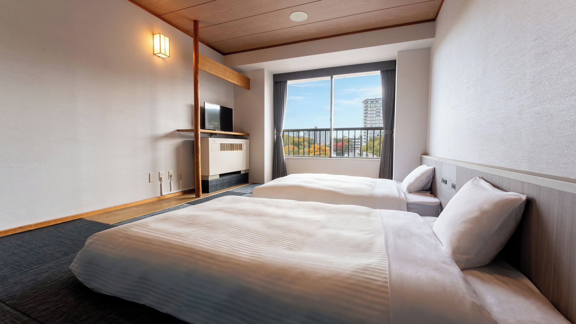 A hotel room with two beds, a TV, a heater, and a window with a view of buildings.