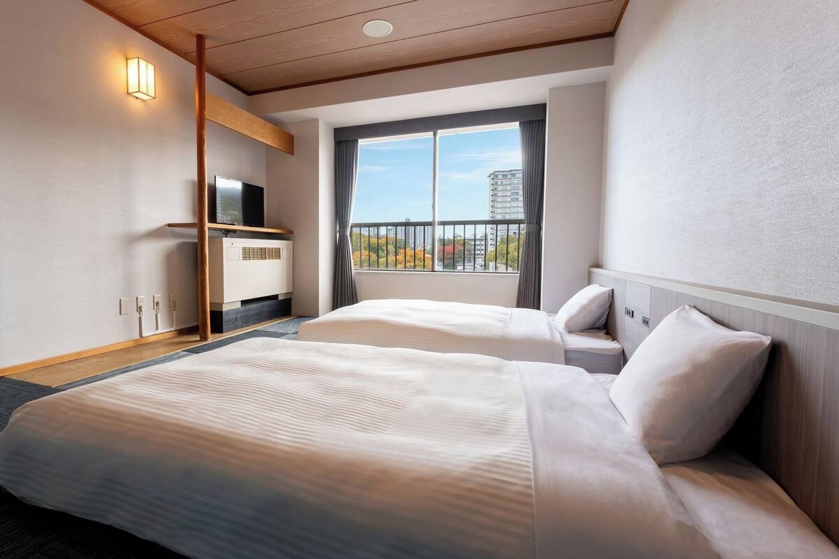Ein Hotelzimmer mit zwei Betten, einem Fernseher, einem Heizkörper und einem Fenster mit Blick auf Gebäude.
