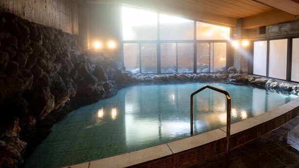 A hot spring bath with a stone wall and a wooden ceiling, surrounded by large windows.