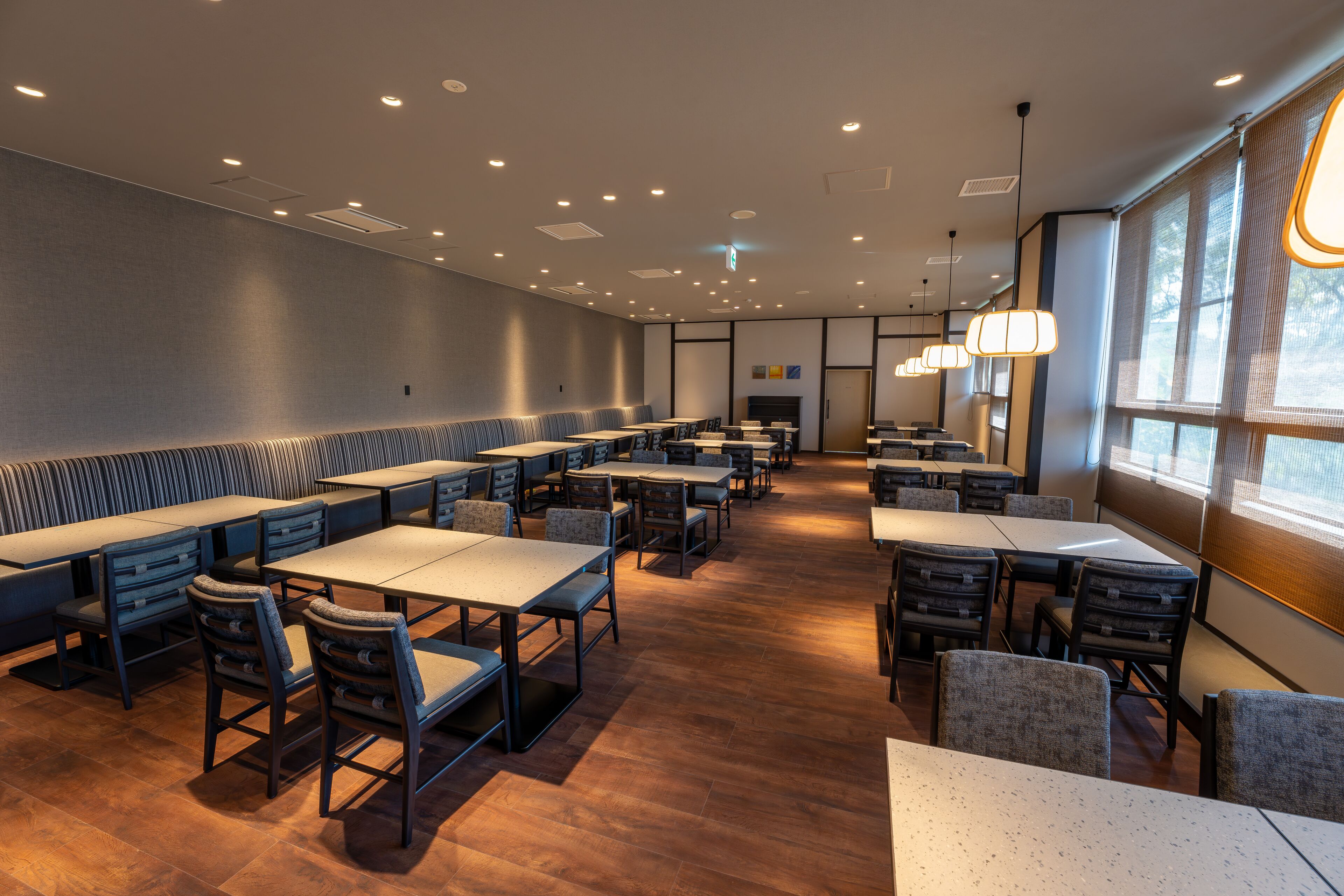 A spacious dining area with long tables and booths, recessed ceiling lighting, and large windows providing natural light.
