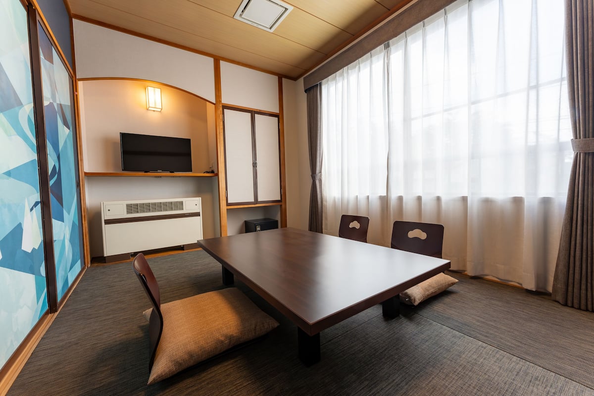 Ein modernes Hotelzimmer mit einem großen Holztisch, bequemen Sitzgelegenheiten, einem Flachbildfernseher und großen Fenstern mit Vorhängen.