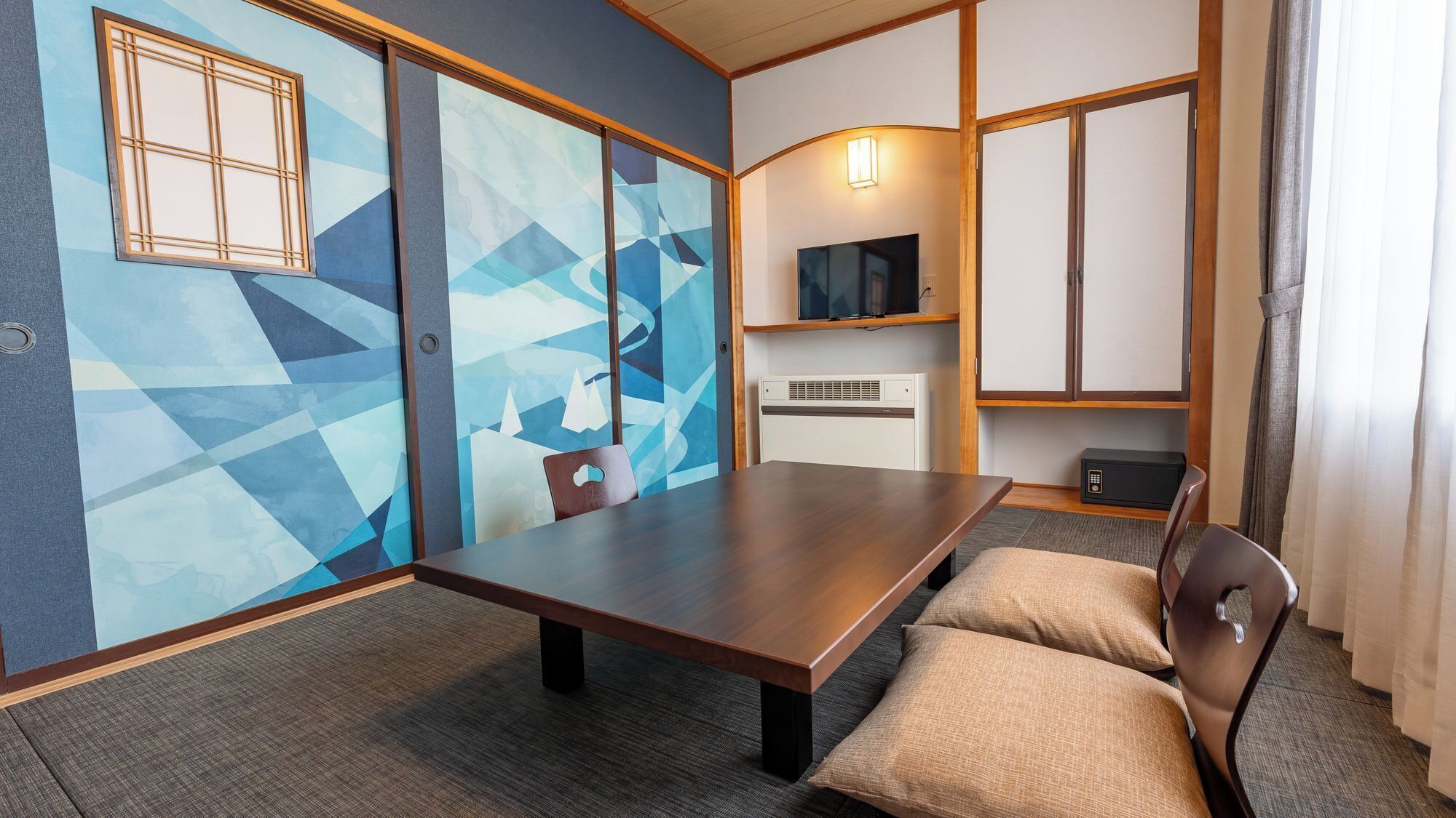 A hotel room with a large wooden table, a TV mounted on the wall, a heater, and a window with curtains.