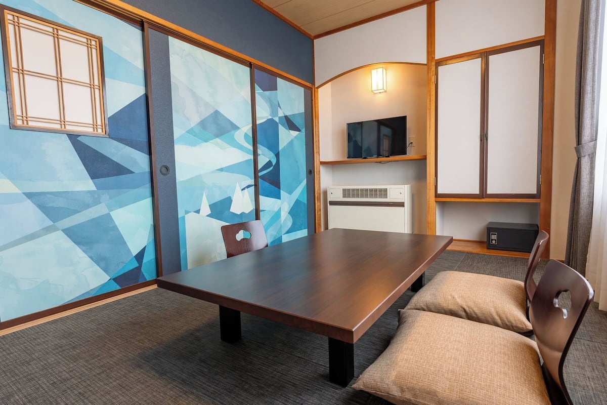 Ein Hotelzimmer mit einem großen Holztisch, einem an der Wand befestigten Fernseher, einem Heizgerät und einem Fenster mit Vorhängen.