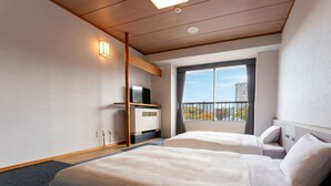 A hotel room with two beds, a TV on a wooden shelf, a window with a view of buildings and trees, and a ceiling with wooden beams.