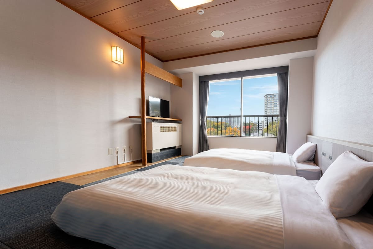 Ein Hotelzimmer mit zwei Betten, einem Fernseher auf einem Holzboden, einem Fenster mit Blick auf Gebäude und Bäume sowie einer Decke mit Holzbalken.