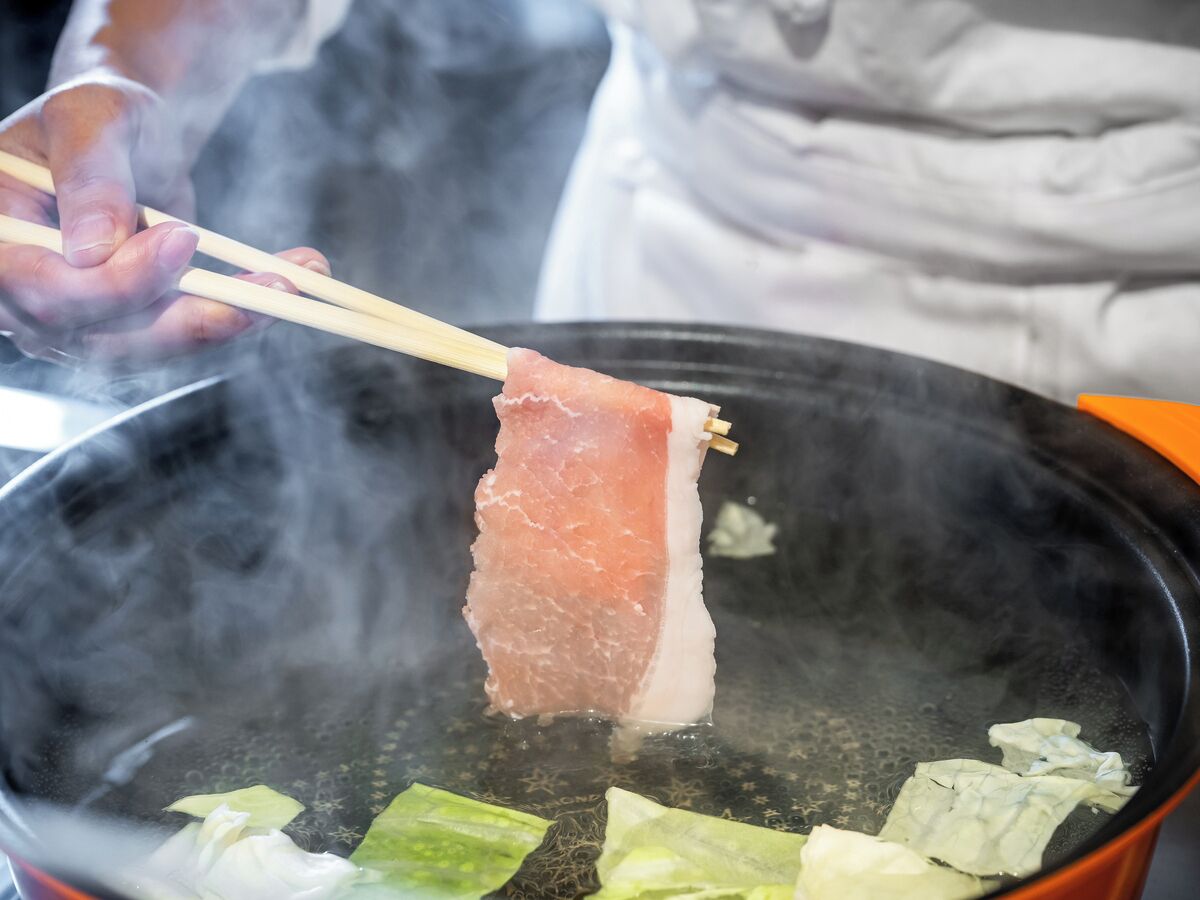 A person holding chopsticks over a pot of boiling water with steam rising, containing pieces of meat and green vegetables.