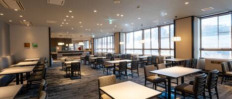 A spacious dining area with multiple tables and chairs, large windows, and ceiling lights.