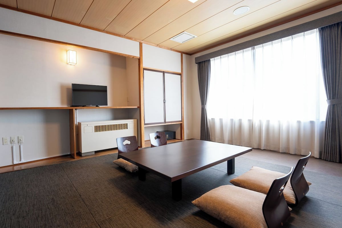 Ein Hotelzimmer mit Holzboden, einem Flachbildfernseher, einem kleinen Heizgerät, einer Couch und einem großen Fenster mit Vorhängen.