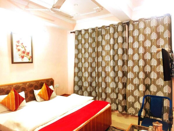 A hotel room with a bed, a red bedspread, patterned curtains, a framed picture on the wall, and a television on the wall. - Kumar Residency (Dharamshala)
