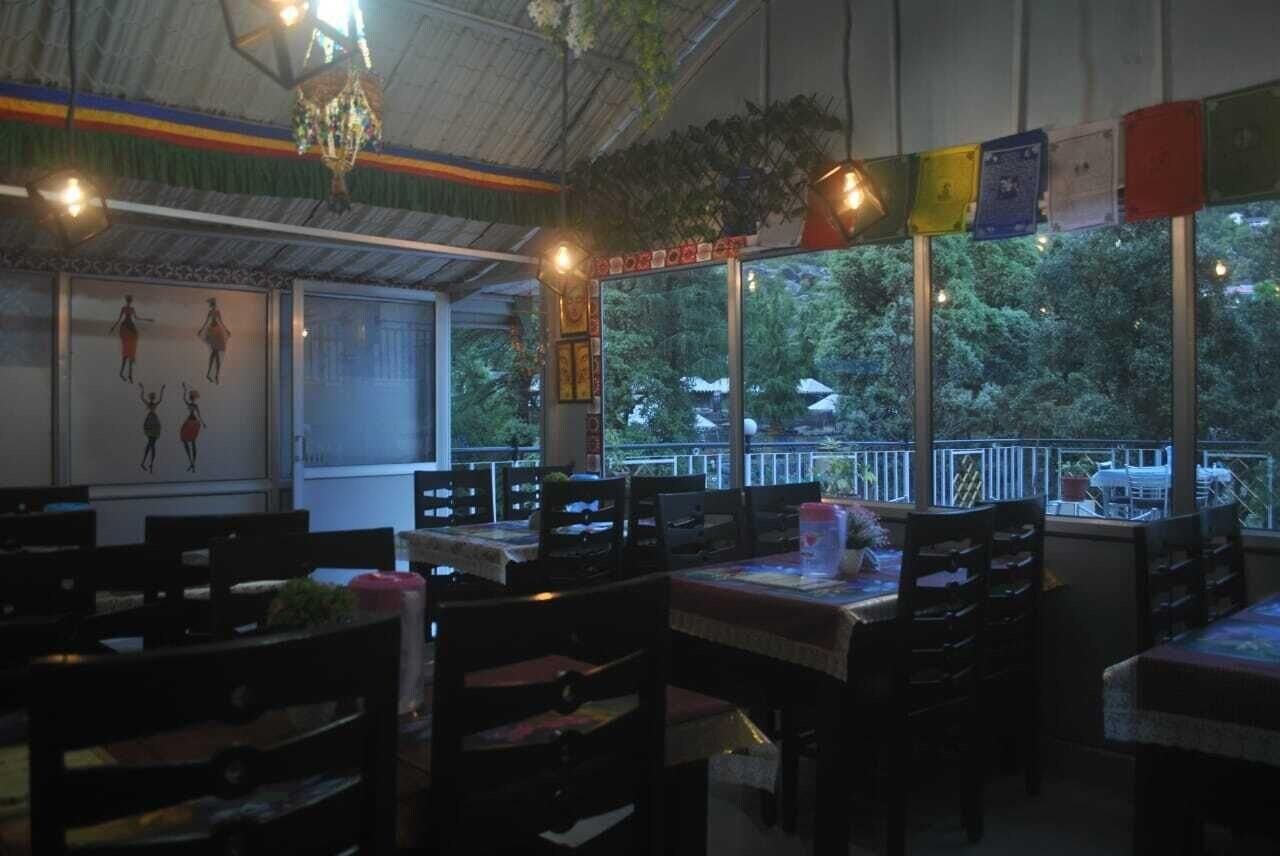 An indoor dining area with tables and chairs, a view through large windows, and a ceiling with hanging lights.