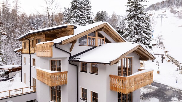Un chalet à plusieurs étages, doté de balcons en bois et de grandes fenêtres, entouré d'arbres couverts de neige et avec un télésiège visible au loin.