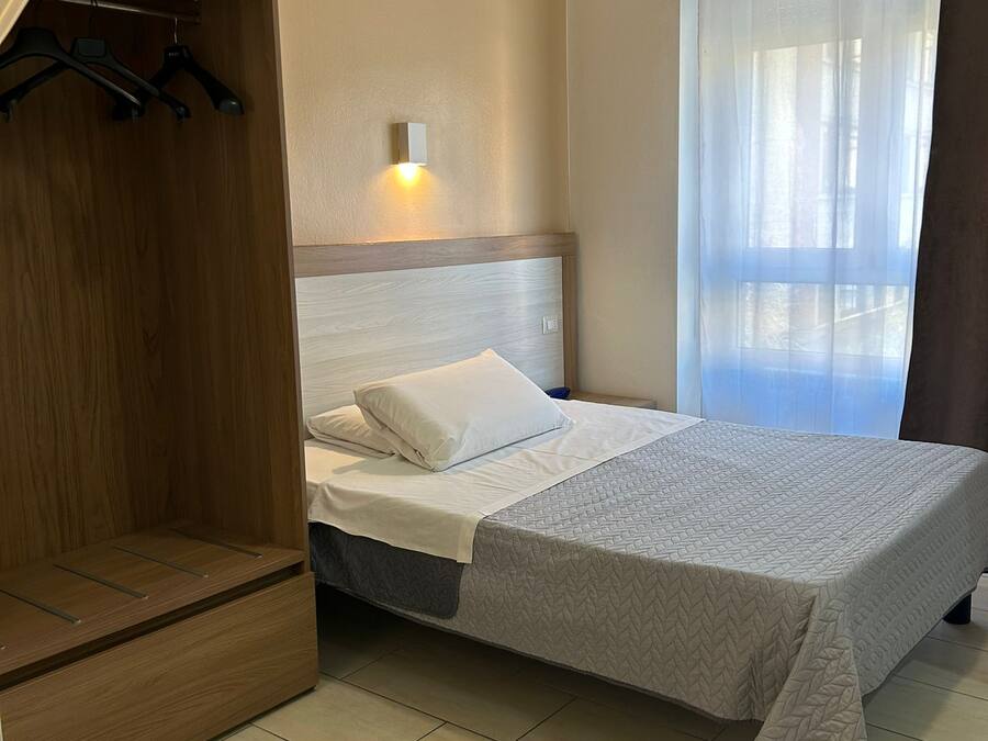 A bedroom with a bed, a wardrobe, a bedside table, a wall-mounted light, and a window with curtains.