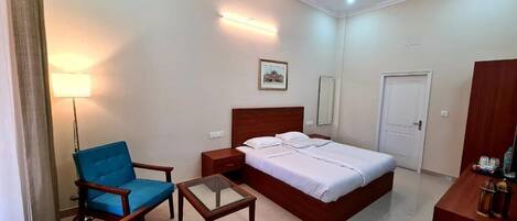A hotel room with a bed, two blue armchairs, a small table, a ceiling fan, and a nightstand.