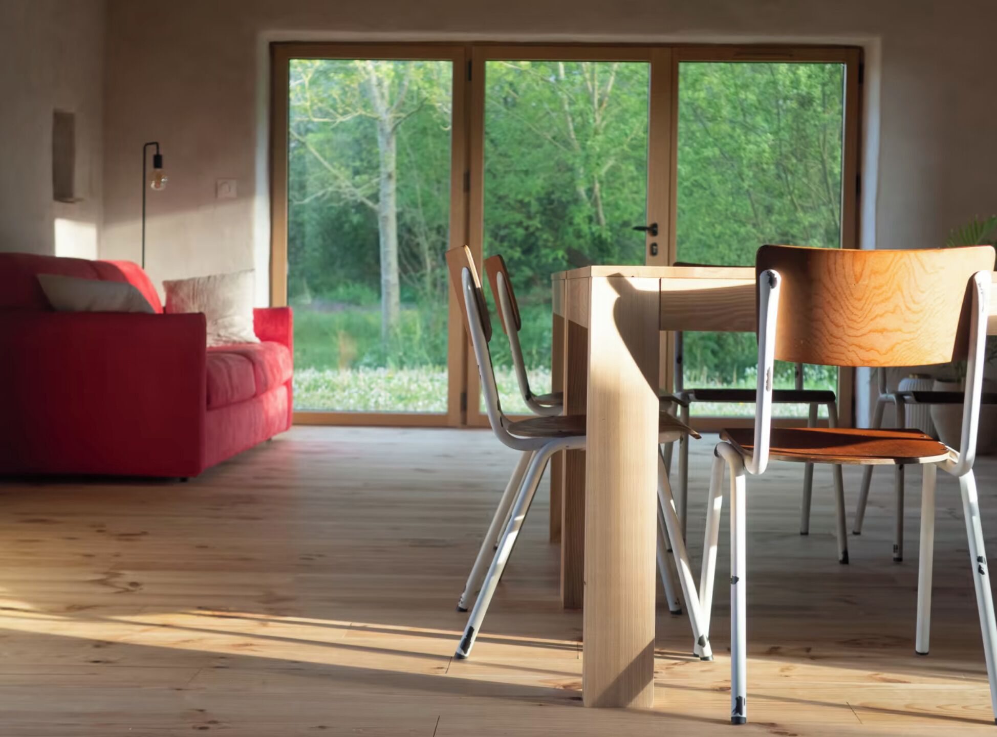 A modern living room with a wooden floor, a red sofa, a dining table with chairs, and a large window with a view of trees.