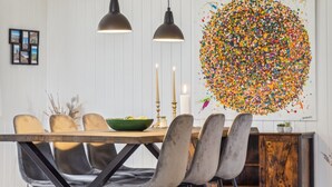 Un comedor con una mesa de madera, sillas negras y lámparas colgantes. Un cuadro abstracto y colorido adorna la pared.