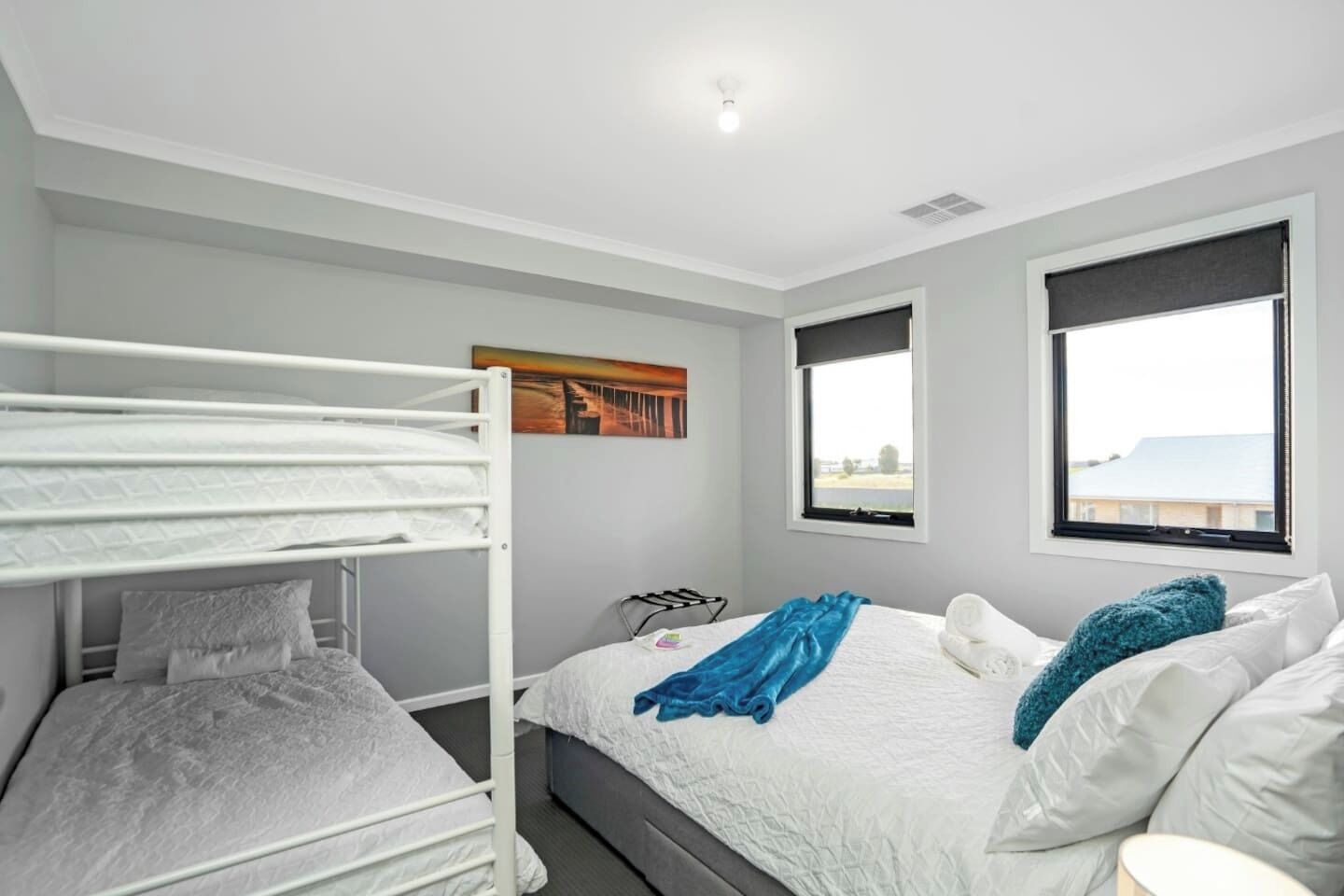 A room with bunk beds, a bed with a blue blanket, a painting on the wall, and a window with a view.