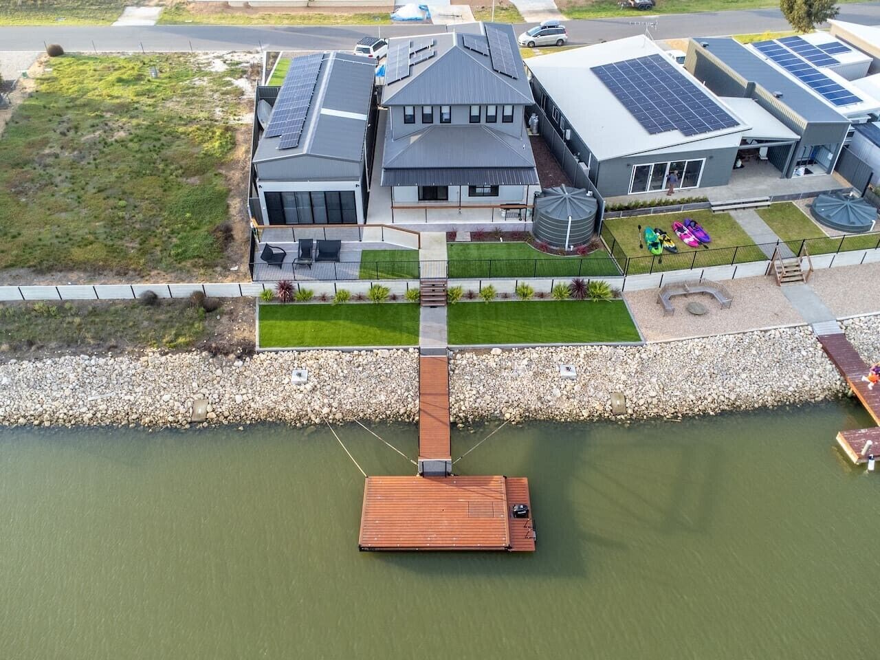 Una casa con paneles solares, un muelle que se adentra en el agua y un espacio verde con un parque infantil.
