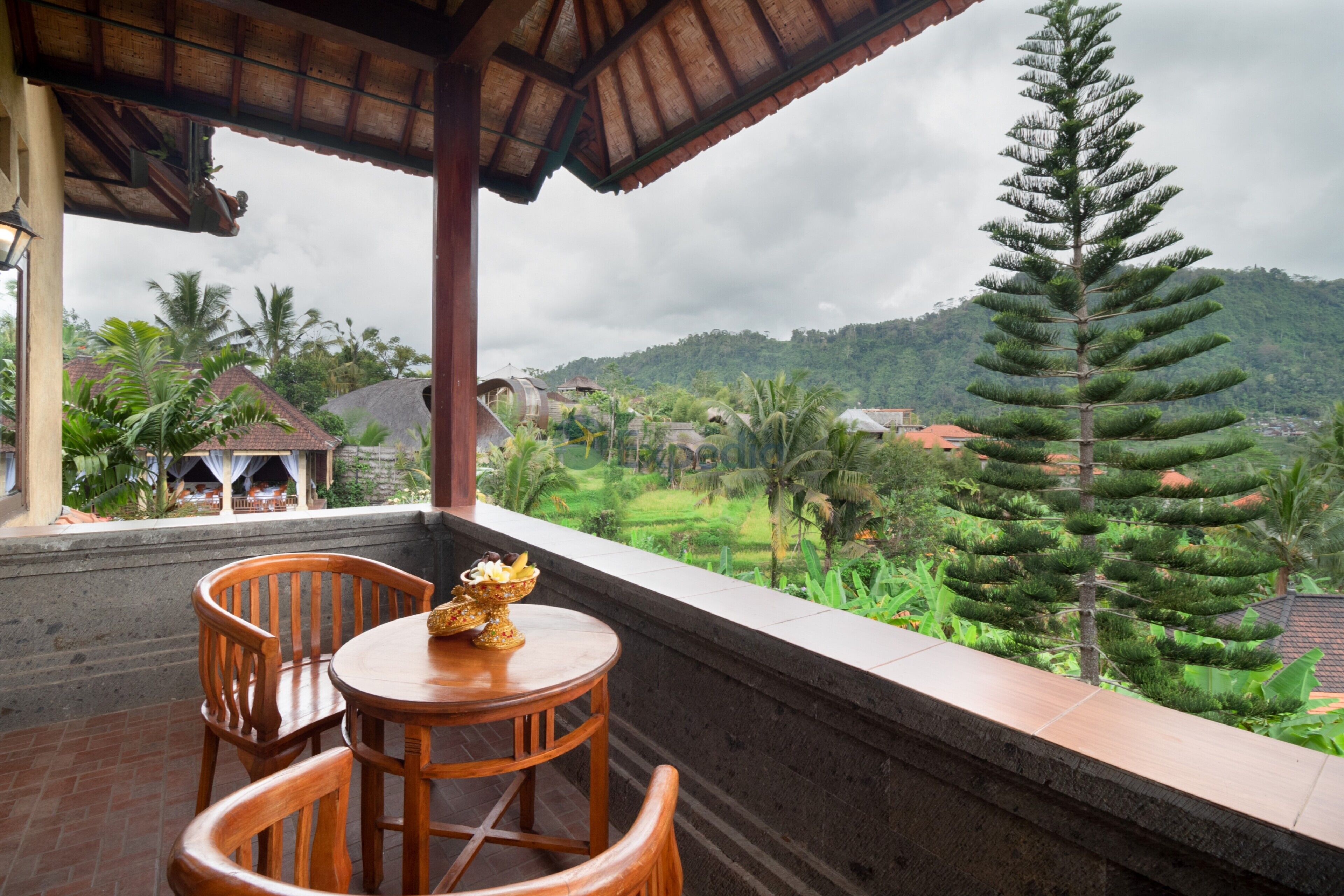 A wooden table and chairs on a balcony with a view of a lush landscape and a tall tree.