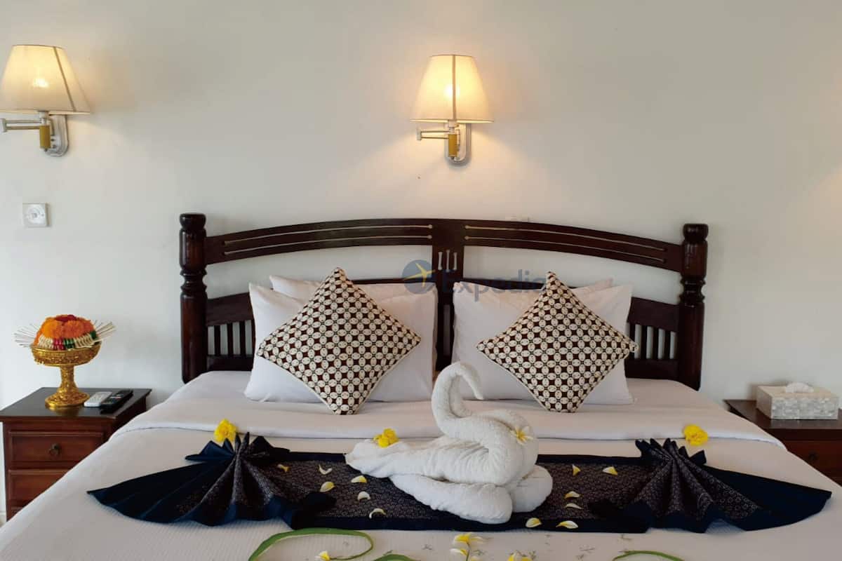 A hotel room with a wooden headboard, two wall-mounted lamps, a bed with white linens, decorative pillows, a swan-shaped towel arrangement, and a small table with a decorative bowl and a phone.