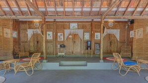 Une pièce en bois avec un plafond haut, deux portes ornées de rideaux décoratifs, des meubles en osier et des tableaux encadrés accrochés aux murs.