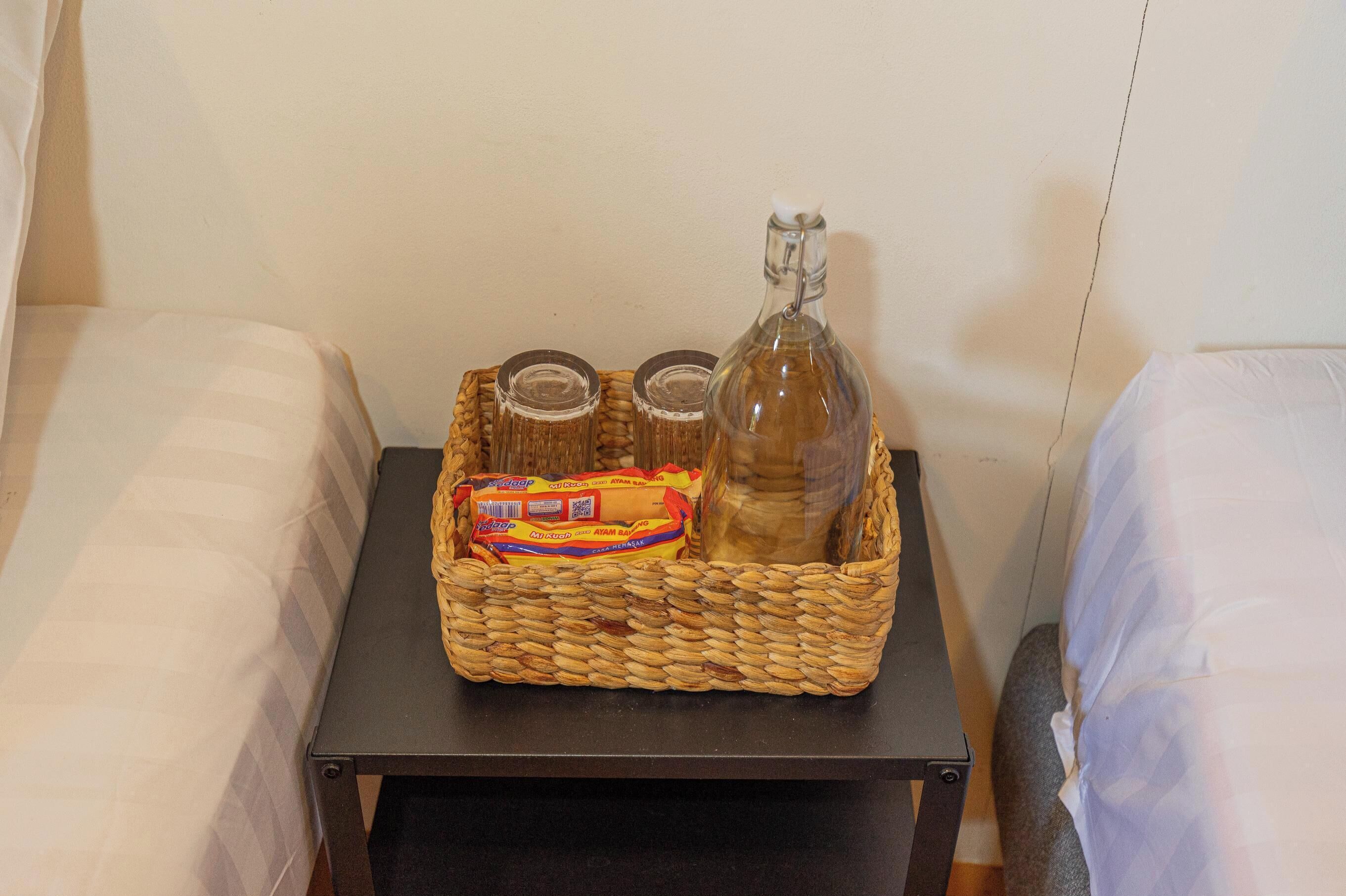 A small table with a woven basket containing a bottle, two glass jars, and a packet of snacks.