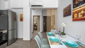 A modern kitchen with a stainless steel refrigerator and oven, a dining area with teal chairs and a white table, and artwork on the walls. - Nice n Cosy Apartment in Rhodes Town (Rodos)