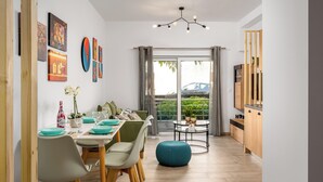 A modern dining area with a table set for four, a blue ottoman, and a kitchen with wooden cabinets and a TV. - Nice n Cosy Apartment in Rhodes Town (Rodos)