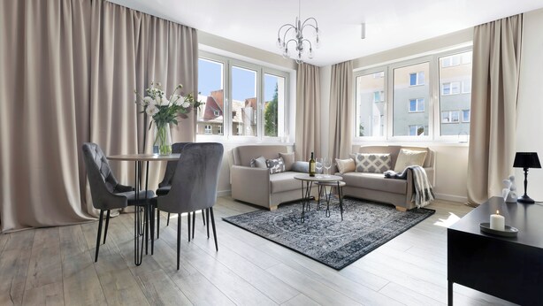 Ein modernes Wohnzimmer mit einer Couch, einem Esstisch und Stühlen. Es gibt eine Chandelüster, eine Vase mit Blumen und ein Fenster mit Vorhängen.