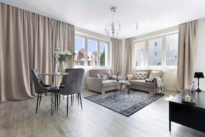 En moderne stue med en sofa, spisebord og stole. Der er en lysekrone, en vase med blomster og et vindue med gardiner.