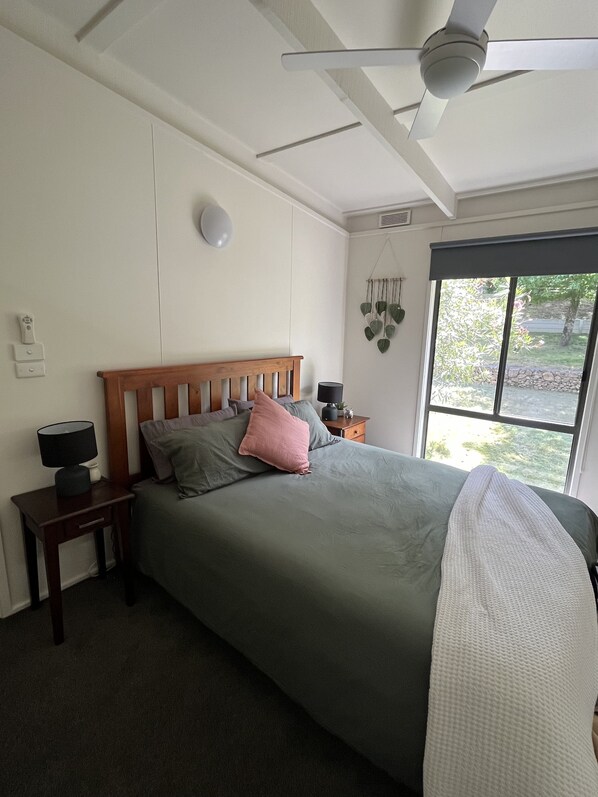 Un dormitorio con cama, mesitas de noche, ventilador de techo, ventana con vista a follaje y una lámpara fijada a la pared.