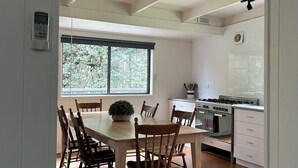 Un coin repas avec une table et des chaises en bois, une plante en pot et une vue sur les arbres à travers une grande fenêtre.