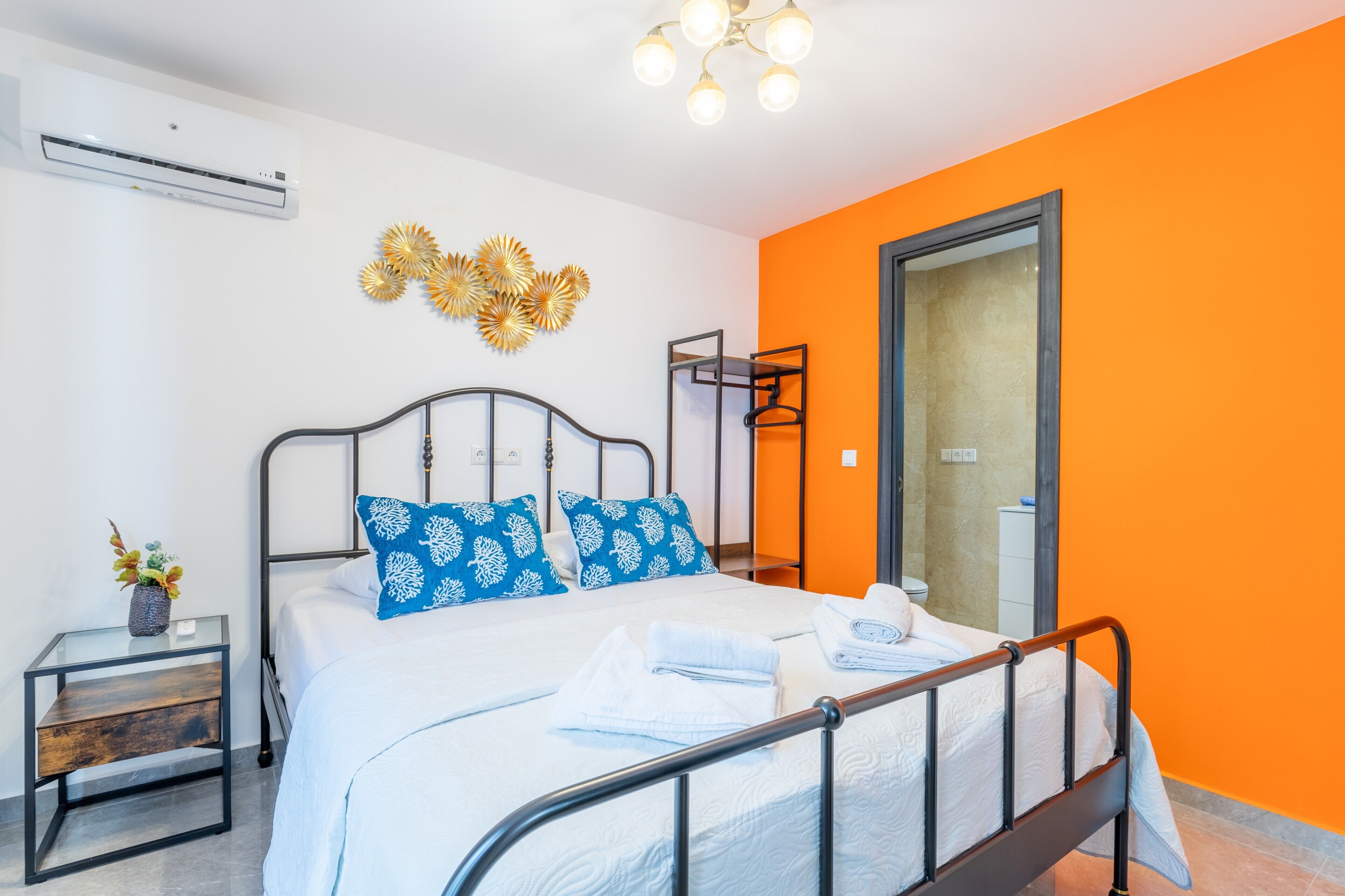 A modern bedroom with a bed, bedside tables, an air conditioner, decorative wall art, and an orange accent wall.